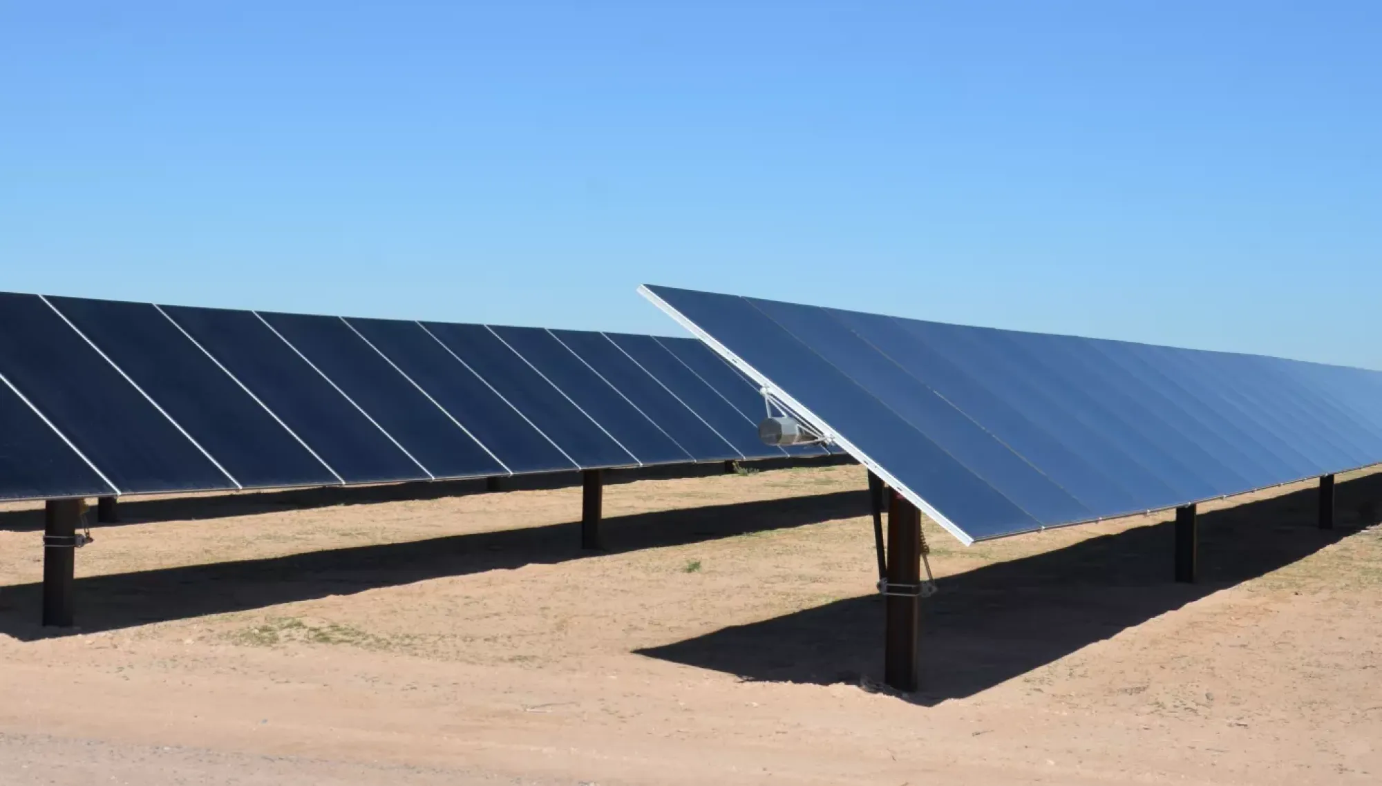 Rows of solar panels on a sandy ground under a clear blue sky.