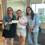 Three women smiling and standing inside a modern office lobby, one holding a large check as they pose for a presentation photo.