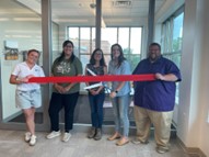Group of five adults holding a red ribbon in front of glass office doors during a ribbon-cutting event, smiling and standing in a hallway.