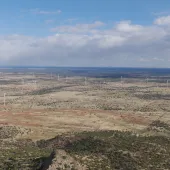 Wind turbines spread across a semi-arid landscape with scattered vegetation under a partly cloudy sky. West Camp 2 wind project.
