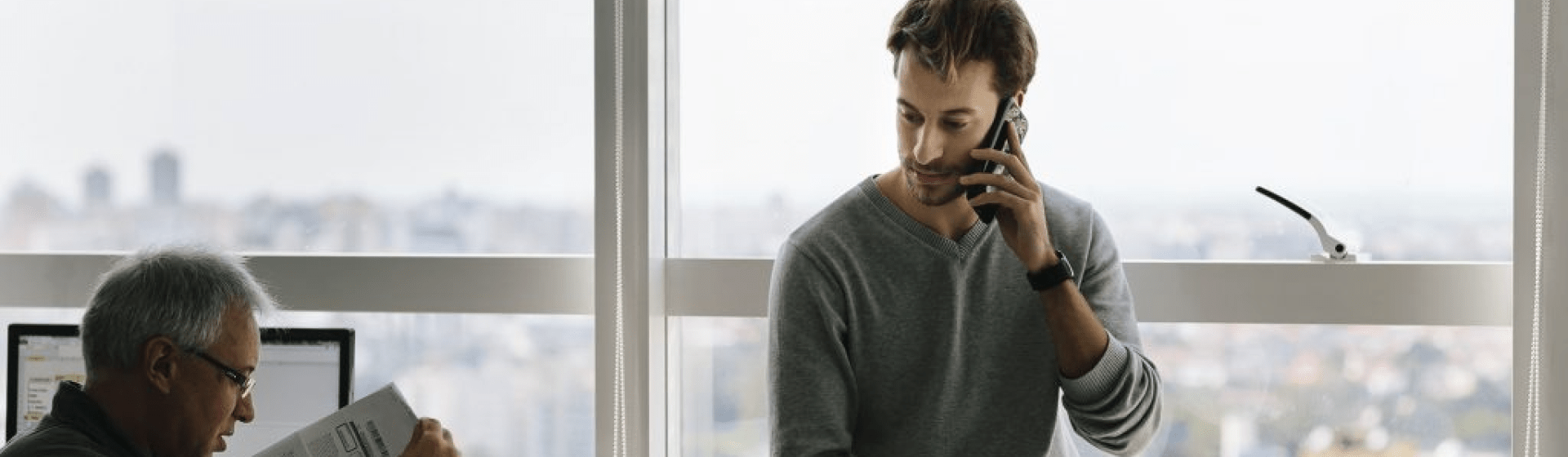 Two men in a windowed office talking; one is sitting on the desk on the phone.