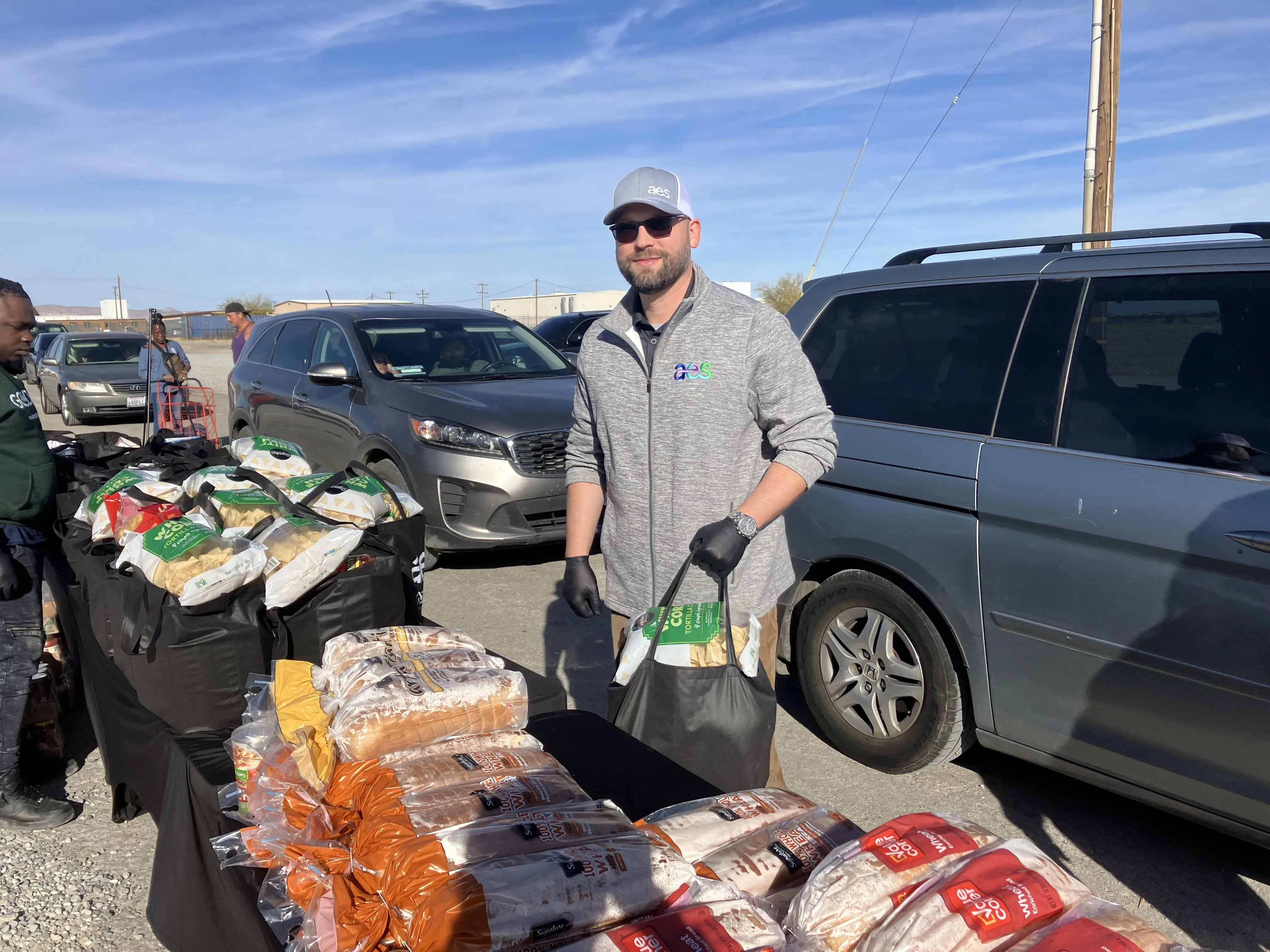 An AES volunteer in sunglasses and gloves stands beside a table filled with bread and bags of groceries, handing out food during a drive-up community distribution event. Vehicles line up in the background as people receive assistance under a sunny blue sky.