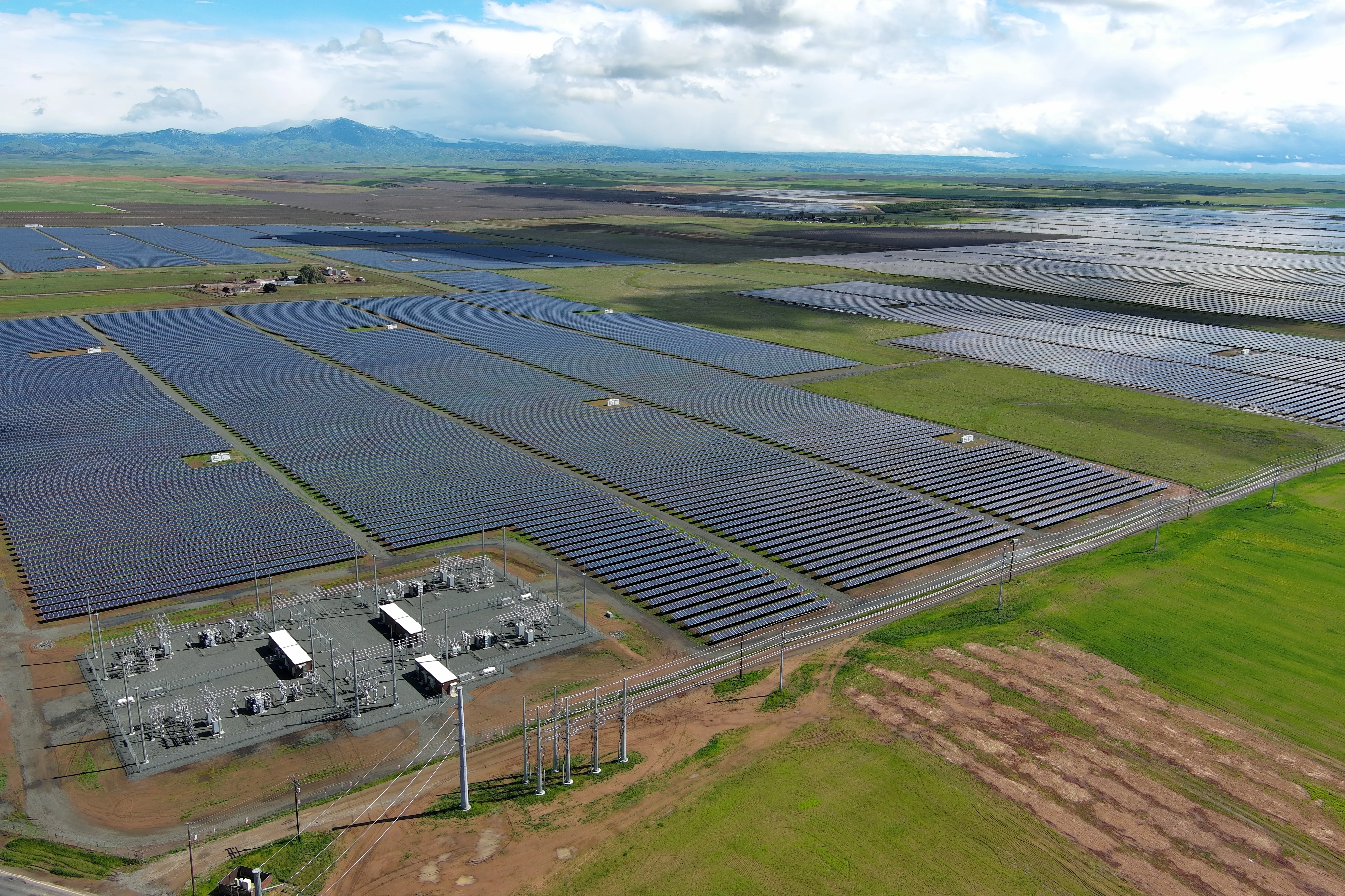 Aerial view of a large-scale solar farm with rows of solar panels stretching across green fields. A power substation is located in the foreground, connected to transmission lines. Rolling hills and distant mountains are visible under a partly cloudy sky.