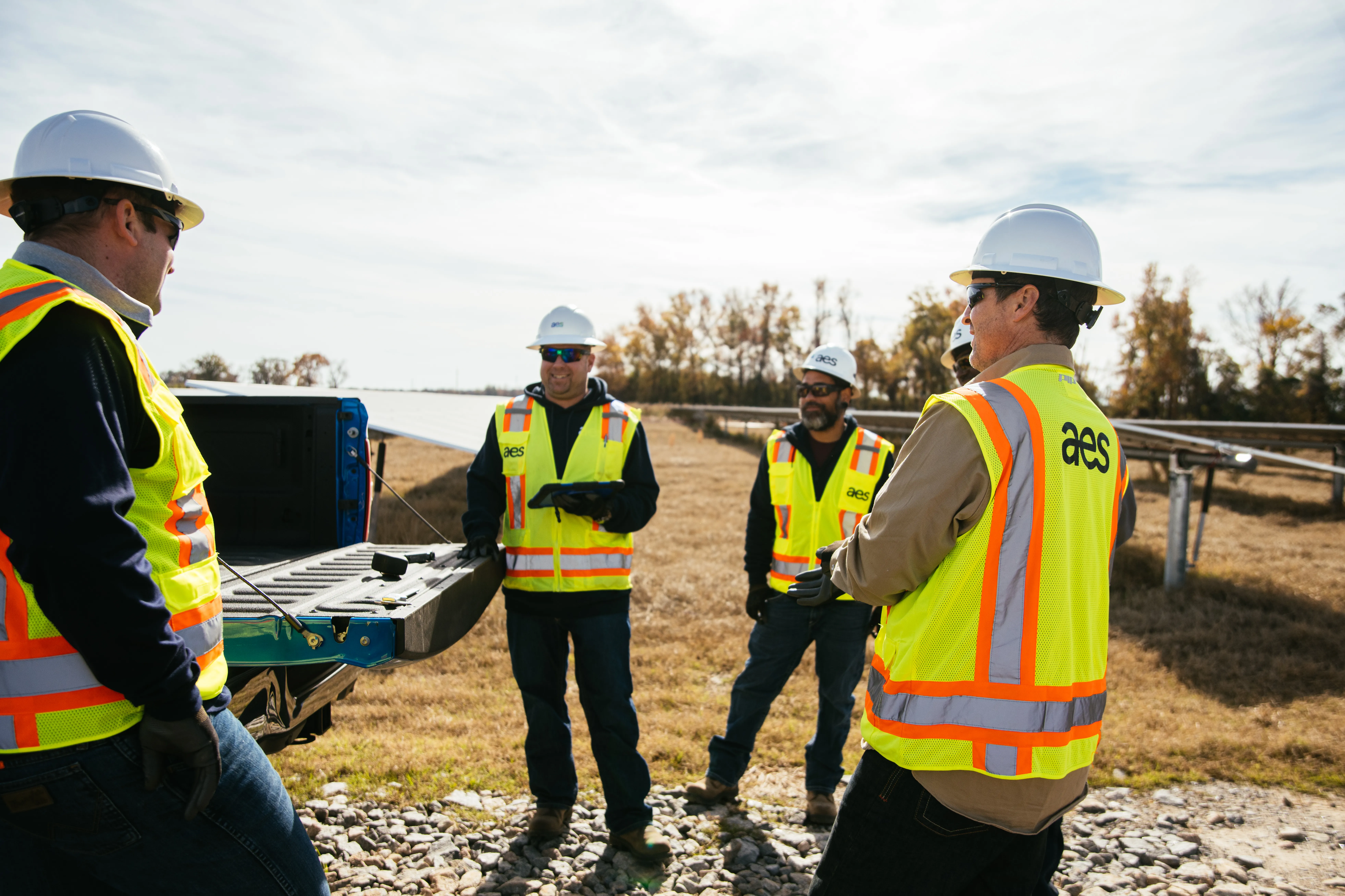 Four AES people wearing high-visibility vests and hard hats conduct a discussion at a solar project site next to a blue pickup truck. Solar panels and autumn trees are visible in the background under a bright sky. The scene conveys teamwork and field safety practices.