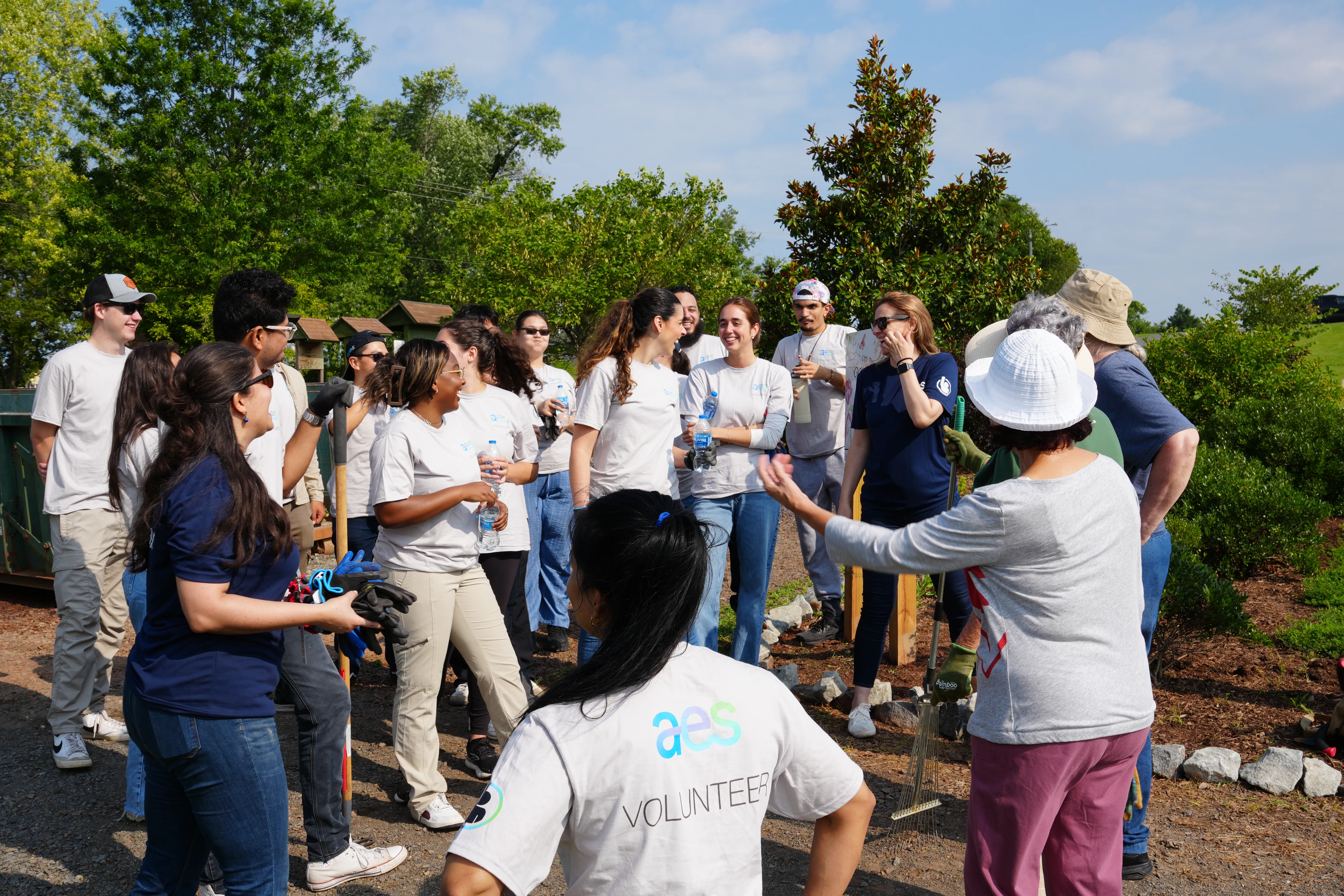 A group of AES volunteers stands together outdoors in a green space with trees and shrubs, preparing for a community service activity. Most people are wearing AES T-shirts and holding tools or water bottles, engaging in conversation under a sunny sky, suggesting a team-oriented event.