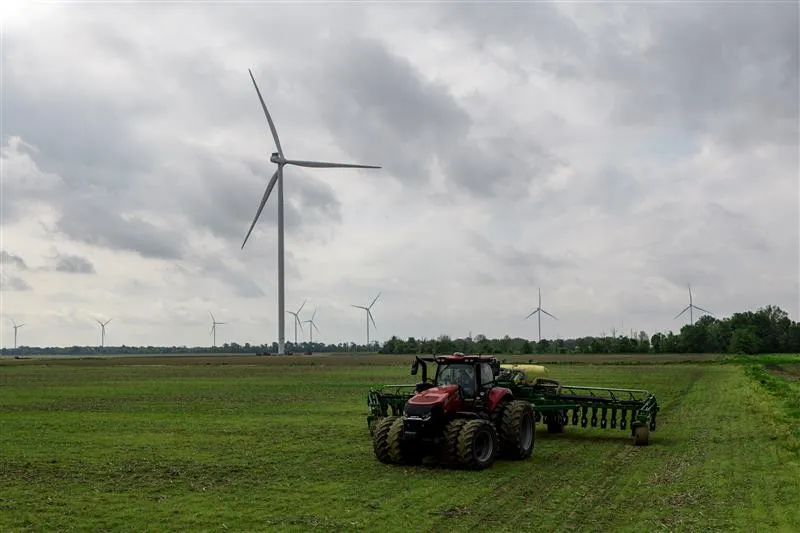 Red tractor in a green farm field with wind turbines in the background under a cloudy sky, highlighting sustainable agriculture and clean energy.