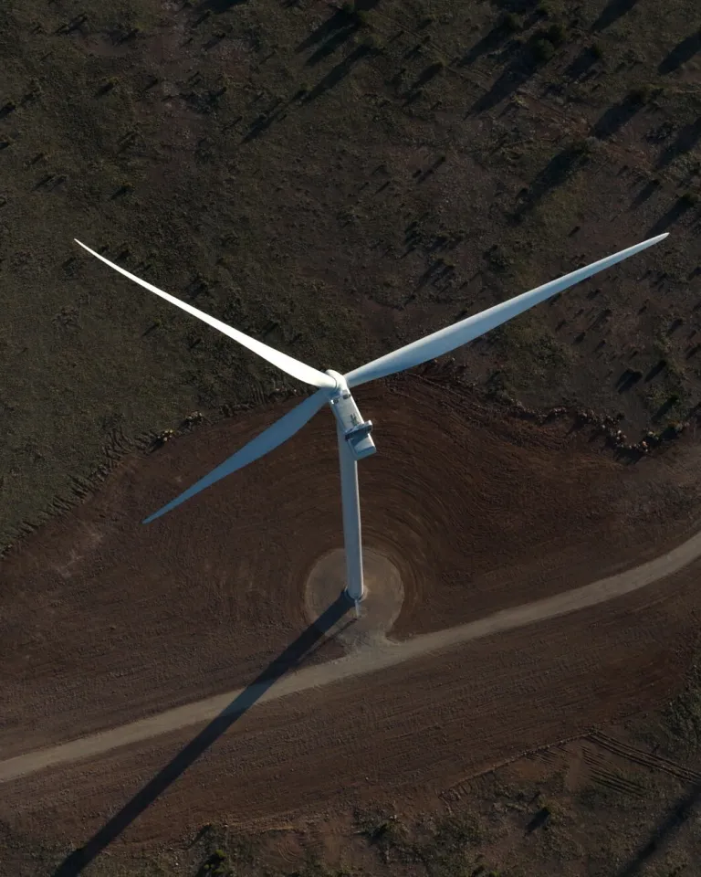 Aerial view of a large wind turbine in a rural area, casting long shadows on the ground. The surrounding landscape is sparsely vegetated with a dirt road leading to the turbine.