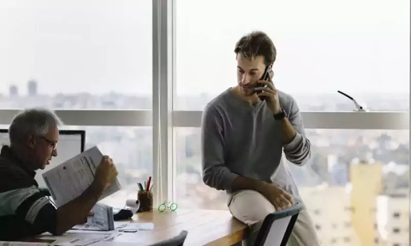 Two men in a windowed office talking; one is sitting on the desk on the phone