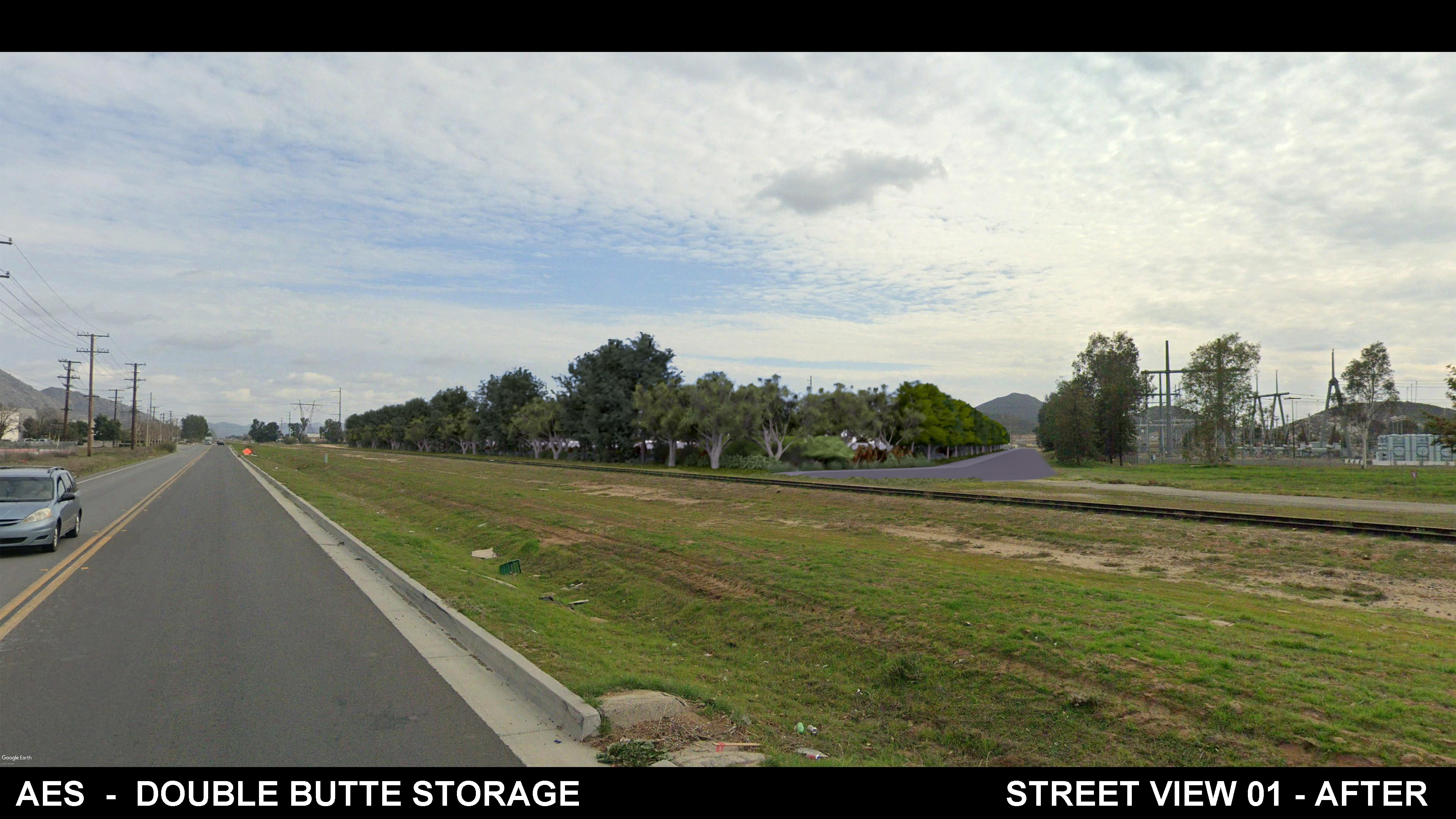 A wide street view shows a road alongside a grassy area and railroad tracks, with a row of mature trees and shrubs newly added along the right side, partially screening an electrical substation and hills in the background under a cloudy sky.