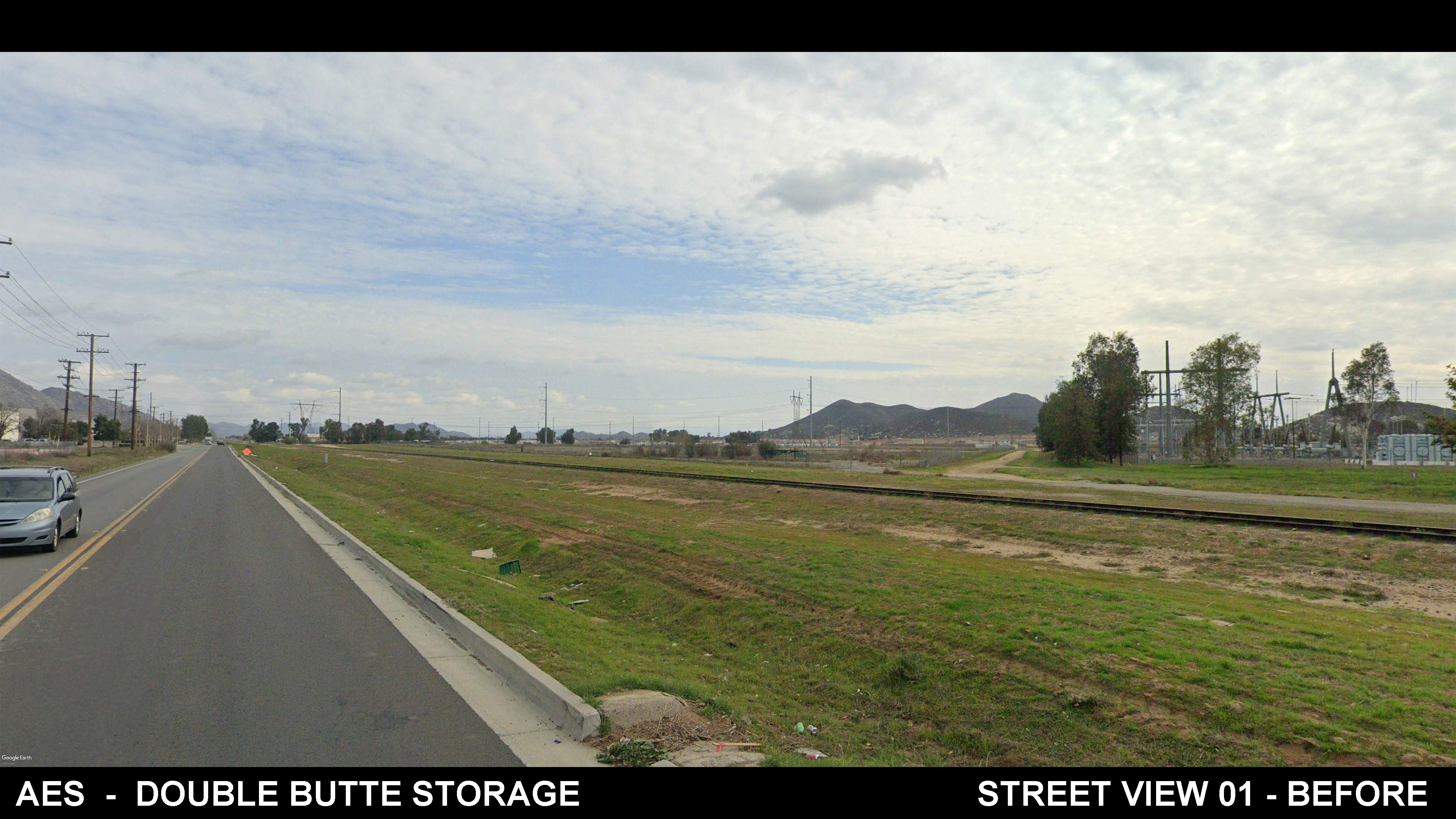 A wide street runs alongside a grassy field with a railroad track, utility poles, and an electrical substation. Mountains and trees are visible in the background under a partly cloudy sky.