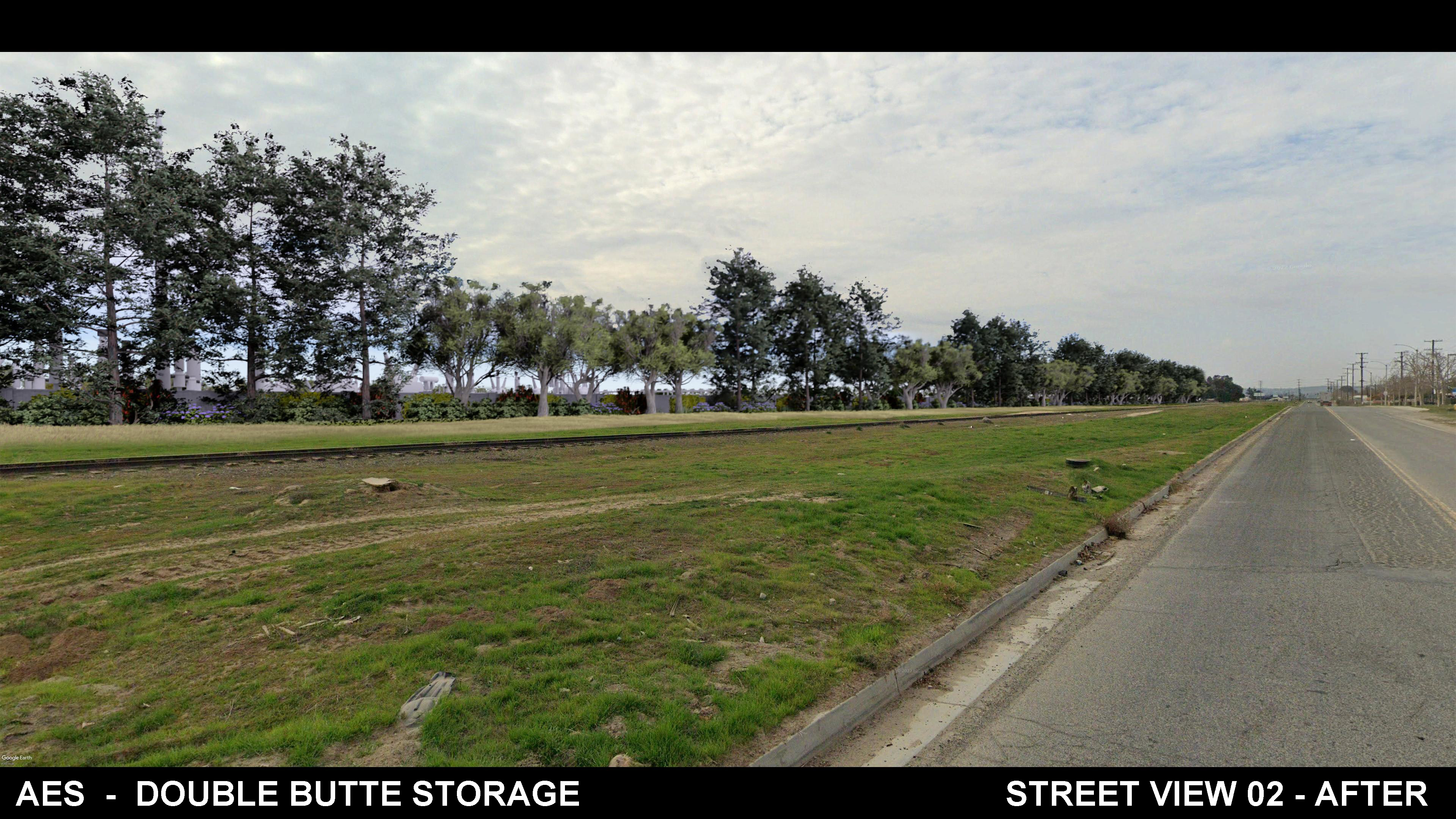 A street view showing a paved road on the right, a grassy area in the center, and a row of tall trees lining the left side. The sky is mostly cloudy, and the area appears clean and well-maintained. Some industrial buildings are partially visible behind the trees.