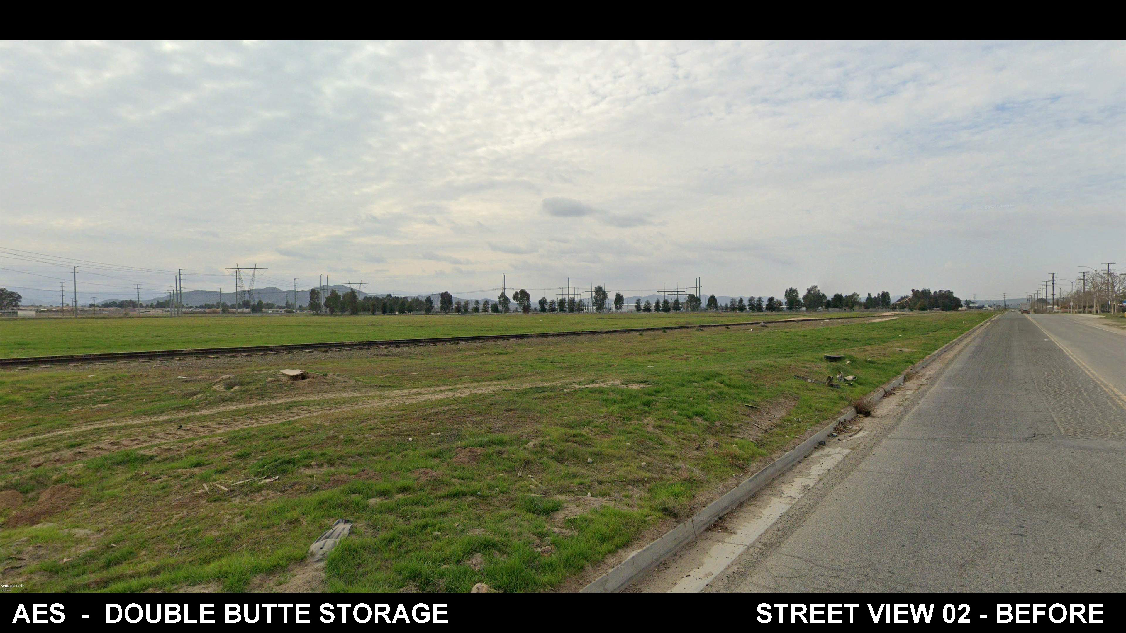A wide view of an empty grassy field with a single railroad track running through it, adjacent to a paved road under a cloudy sky. Power lines and distant trees are visible in the background.