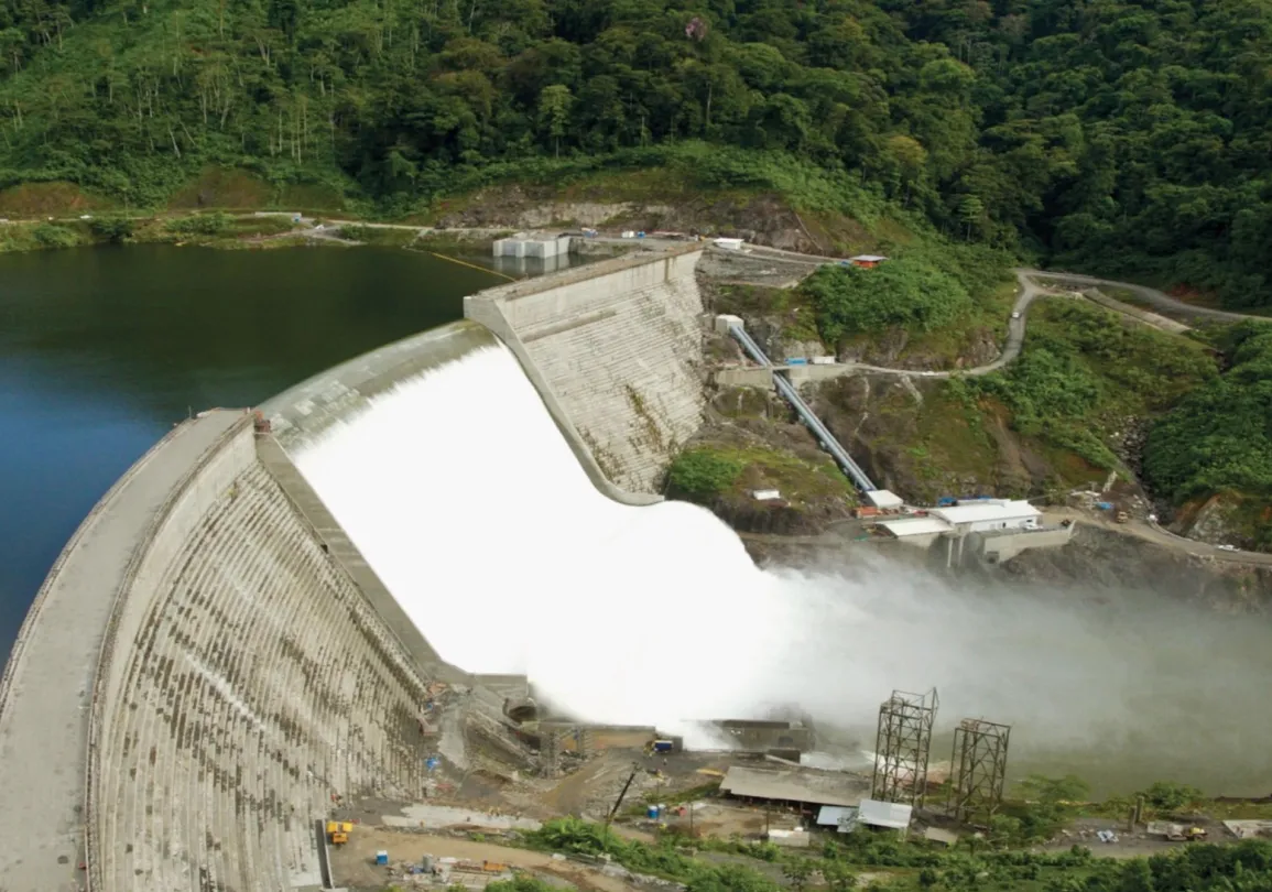 Aerial view of a large dam with water flowing over it, surrounded by lush green forest and a winding road.