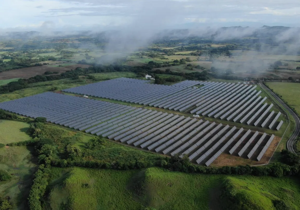 Aerial view of a large solar farm with rows of solar panels surrounded by lush green fields and distant hills under a cloudy sky.
