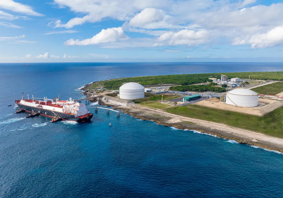 Aerial view of a coastal industrial area with two large white storage tanks near the shoreline. A large ship is docked at the pier, surrounded by clear blue water. The background shows green vegetation and a blue sky with scattered clouds.