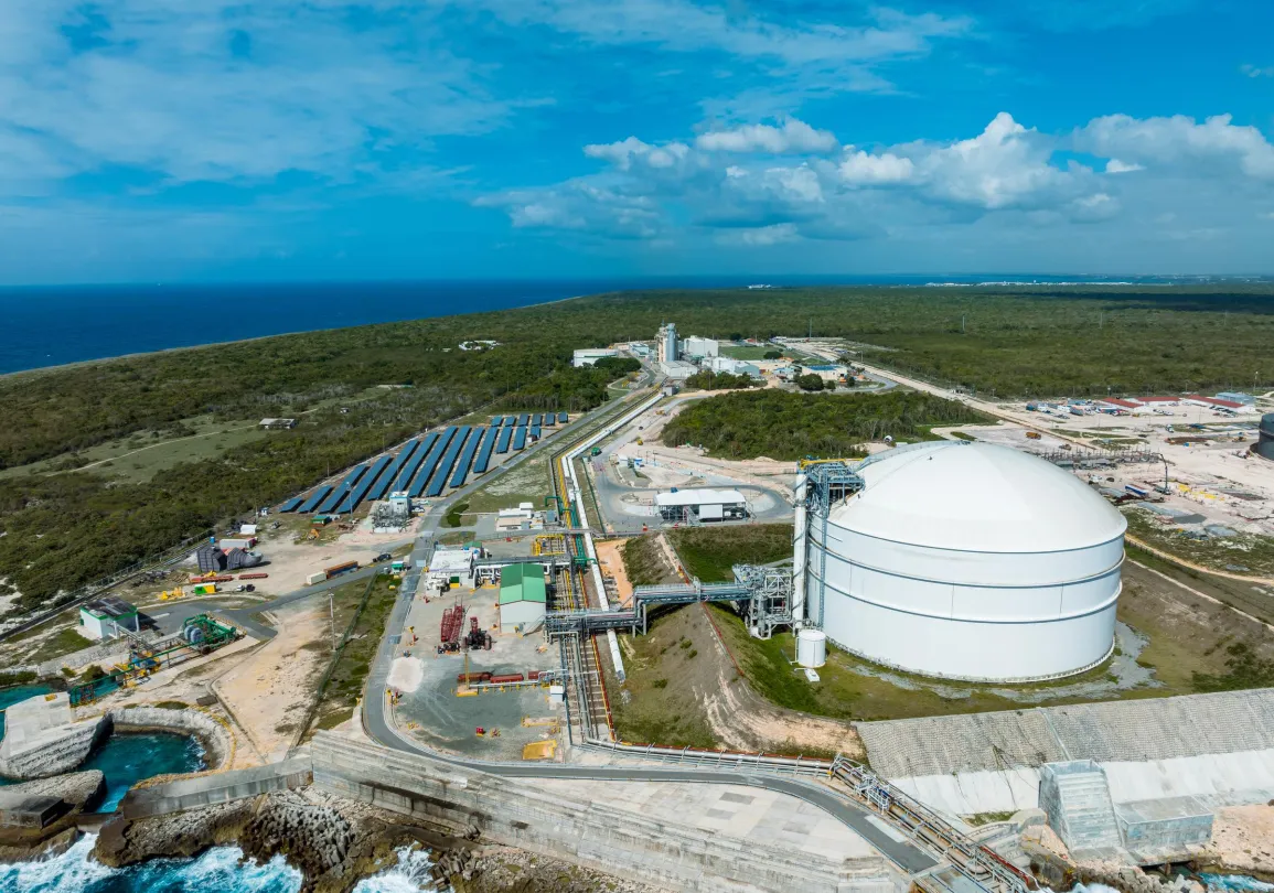 Aerial view of an industrial complex near the coast, featuring a large white storage tank, solar panels, and various buildings surrounded by greenery. The ocean is visible in the background under a partly cloudy blue sky.