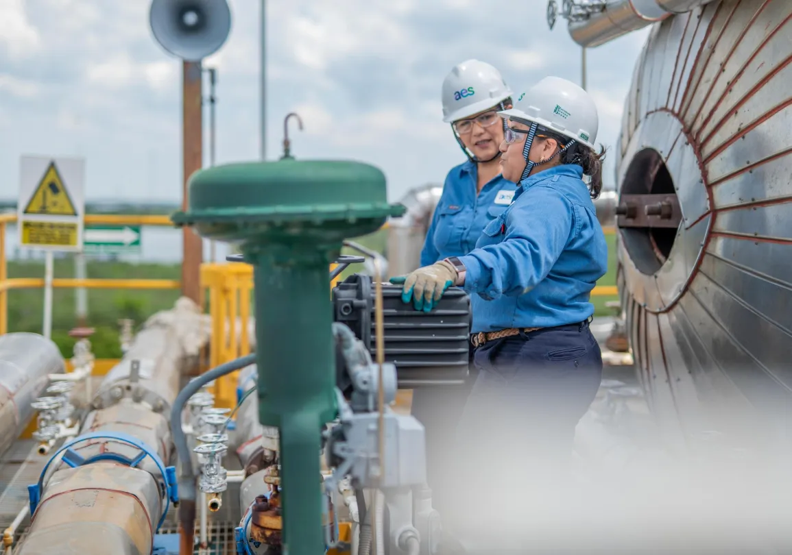 Two workers in blue uniforms and white helmets inspect industrial equipment outdoors. They stand near large metal pipes and a cylindrical tank. One worker points at a device while the other observes. Safety signs and a loudspeaker are visible in the background under a cloudy sky.