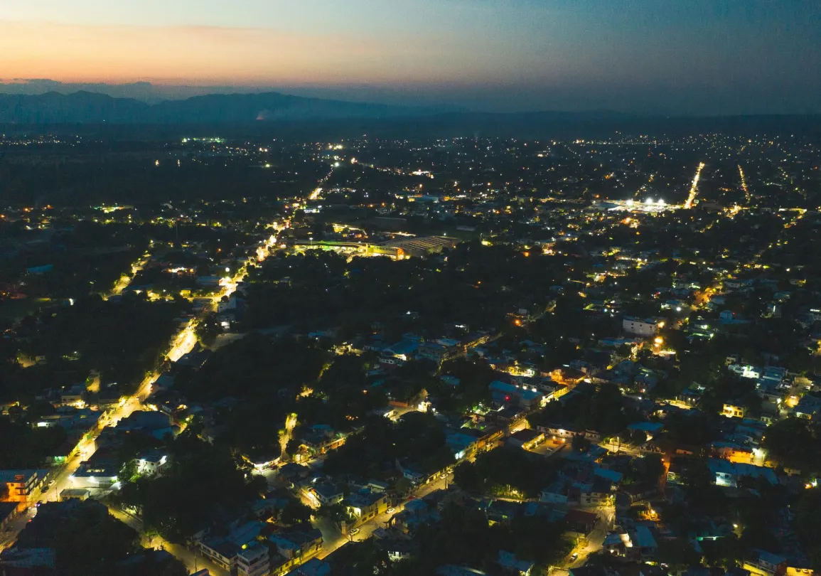 Aerial view of a city at dusk with illuminated streets and buildings, surrounded by darkened landscape and distant mountains under a fading sunset sky.
