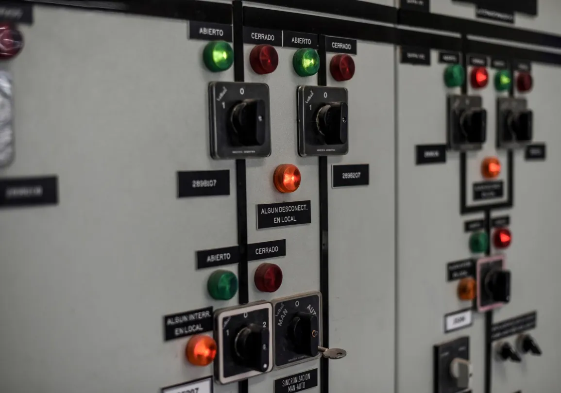 Close-up of an industrial control panel with switches, dials, and indicator lights labeled in Spanish. The panel includes green and red lights indicating open and closed statuses, and various switches with numerical settings.