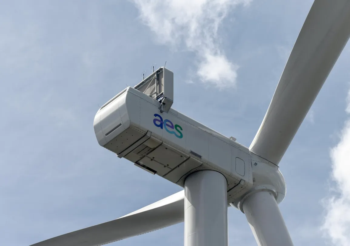 Close-up view of a large wind turbine against a blue sky with clouds. The turbine has the letters 'aes' on its side.