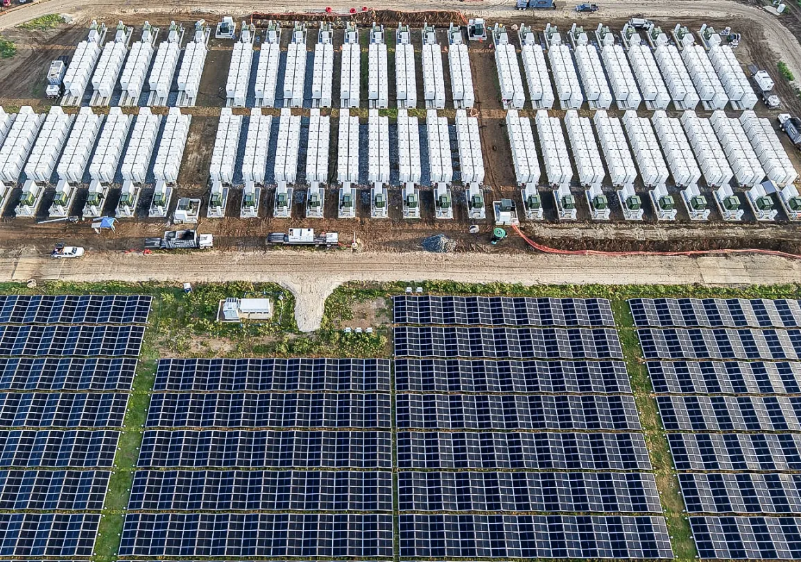 Aerial view of a large solar panel farm next to rows of white battery energy storage system units.