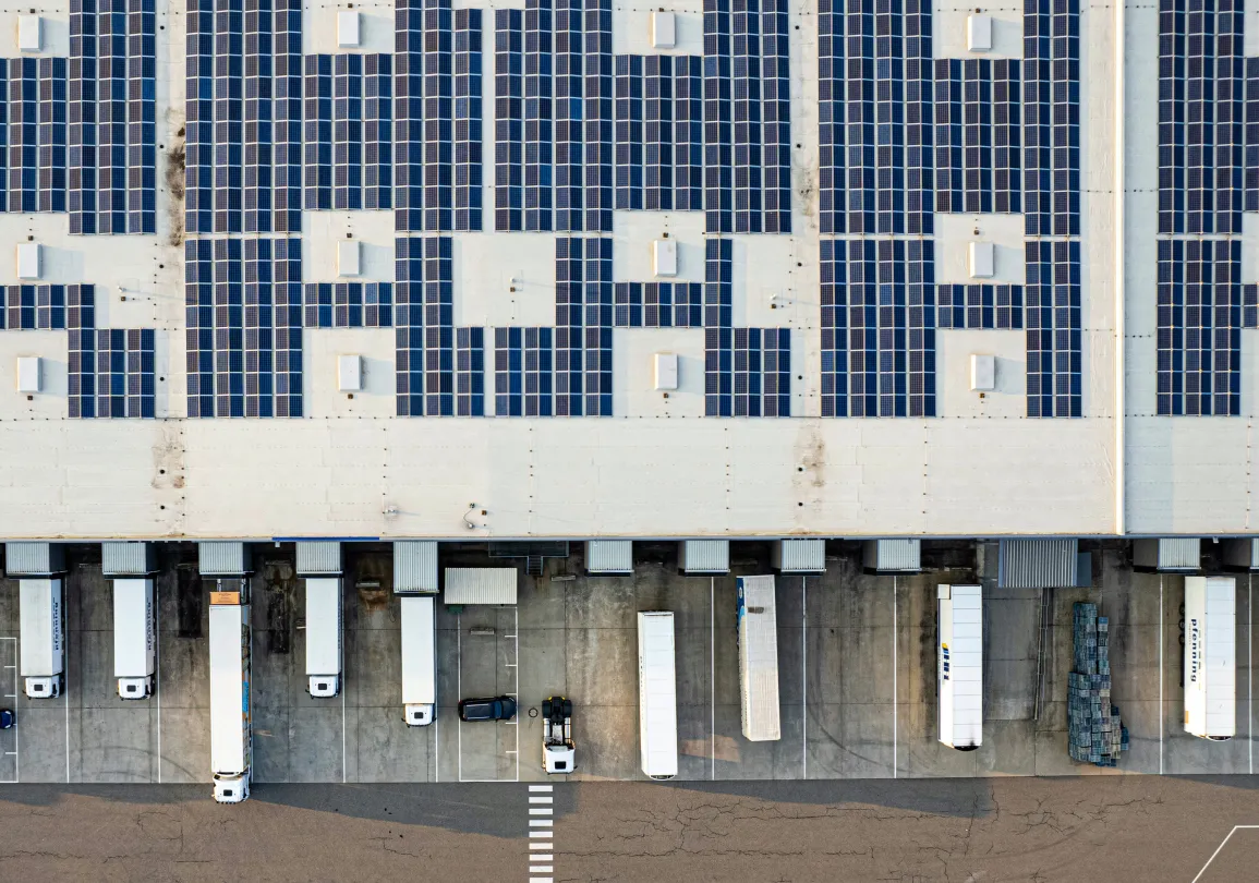 Aerial view of a warehouse rooftop covered in solar panels with several semi-truck trailers parked at the loading docks.