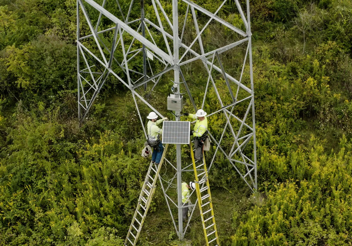 Utility workers on ladders installing a solar panel on a metal transmission tower in a field.