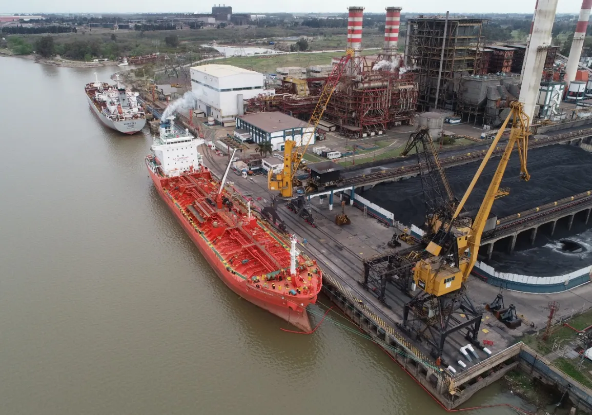 Aerial view of a red cargo ship docked at an industrial port next to large coal piles and a power plant.