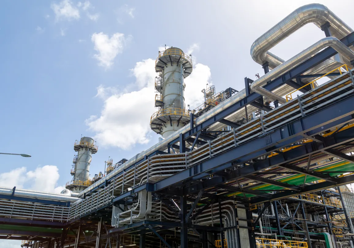 Large industrial distillation towers and complex steel piping systems against a bright blue sky with white clouds.