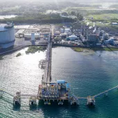 Aerial view of an industrial facility by the water, featuring a large circular storage tank, multiple buildings, and pipelines extending over the water. The facility is surrounded by greenery and has a dock with equipment extending into the sea.