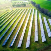 Aerial view of a solar farm with multiple rows of solar panels on a grassy field, surrounded by green countryside and trees.