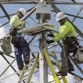 Two utility workers in safety gear and helmets are installing equipment on a metal tower. They are using ladders and are surrounded by a framework of steel beams against a blue sky.