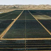 Aerial view of an extensive solar farm with rows of solar panels stretching into the distance, set against a backdrop of arid hills and a clear sky.