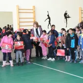 A group of children and a few adults stand in a gymnasium holding red gift boxes. The children are wearing winter clothing. There are wall bars and silhouettes of dancers on the wall behind them. A large mirror reflects some of the group.