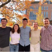 Five people standing together outdoors, smiling under a tree with autumn leaves. A brick building and mountains are visible in the background.