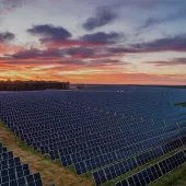 A vast field of solar panels under a vibrant sunset sky with red, orange, and purple hues.