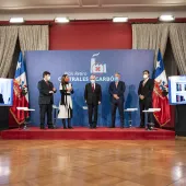 Five people wearing masks stand on a stage in front of a backdrop with the text 'Plan Retiro Centrales Carbón.' Two screens on either side display virtual participants. The setting is formal, with flags and curtains in the background.