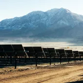 Rows of solar panels on a dirt field with snow-capped mountains in the background under a clear blue sky.