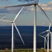 A row of wind turbines in a vast open landscape under a partly cloudy sky. The turbines are large with white blades, and the view extends to a distant horizon.