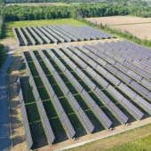 Aerial view of a large solar farm with multiple rows of solar panels set in a grassy field. The panels are aligned in parallel rows, surrounded by green trees and open fields in the background.