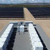 Aerial view of a solar farm with rows of solar panels and white utility boxes on a gravel path, set in a desert landscape under a cloudy sky.