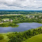 Aerial view of a large solar panel farm surrounded by green fields and trees. In the background, there are residential areas with houses and roads, set against a backdrop of rolling hills and a cloudy sky.