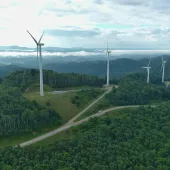 Aerial view of a lush green mountain landscape with multiple wind turbines on hilltops under a partly cloudy sky.
