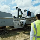 A person wearing a safety vest and hard hat observes a solar panel installation robot labeled 'maximo' on a sunny day.