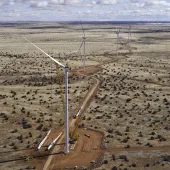 Aerial view of a wind farm under construction in a vast, arid landscape. Several wind turbines are visible, with one turbine being assembled by cranes. A dirt road winds through the sparse vegetation.