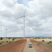 A white truck drives down a dirt road flanked by large wind turbines in a vast, open landscape under a cloudy sky.