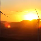 Two wind turbines silhouetted against a vibrant sunset with mountains in the background. The sky is filled with warm hues of orange and yellow, and the sun is partially visible behind the hills.