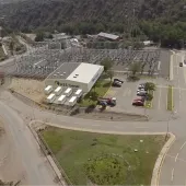 Aerial view of an industrial facility labeled 'AES Gener' surrounded by roads and greenery. The facility includes a main building, parking lot with several cars, and adjacent structures. Nearby, there is a substation with electrical equipment and power lines. The area is set in a mountainous landscape with trees and winding roads.