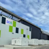 A modern industrial building with a geometric design and the letters 'aes' on the facade. The building features a mix of white, green, and dark panels, with several electrical units in front. The sky is partly cloudy.