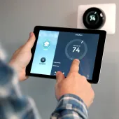 Person adjusting a smart thermostat using a tablet. The screen shows a temperature setting of 74 degrees for the living room. A wall-mounted thermostat displays the same temperature.